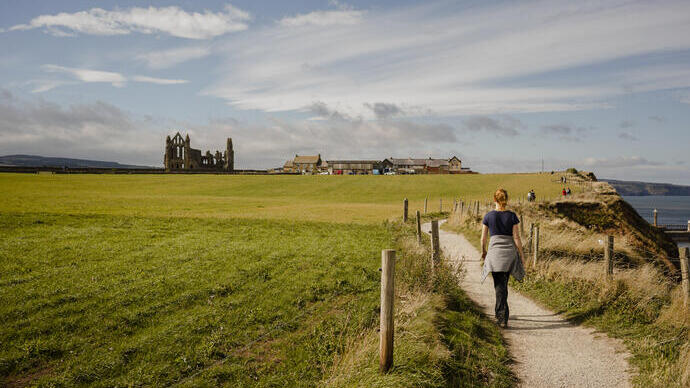 Woman walking along a coastal path towards a ruined abbey