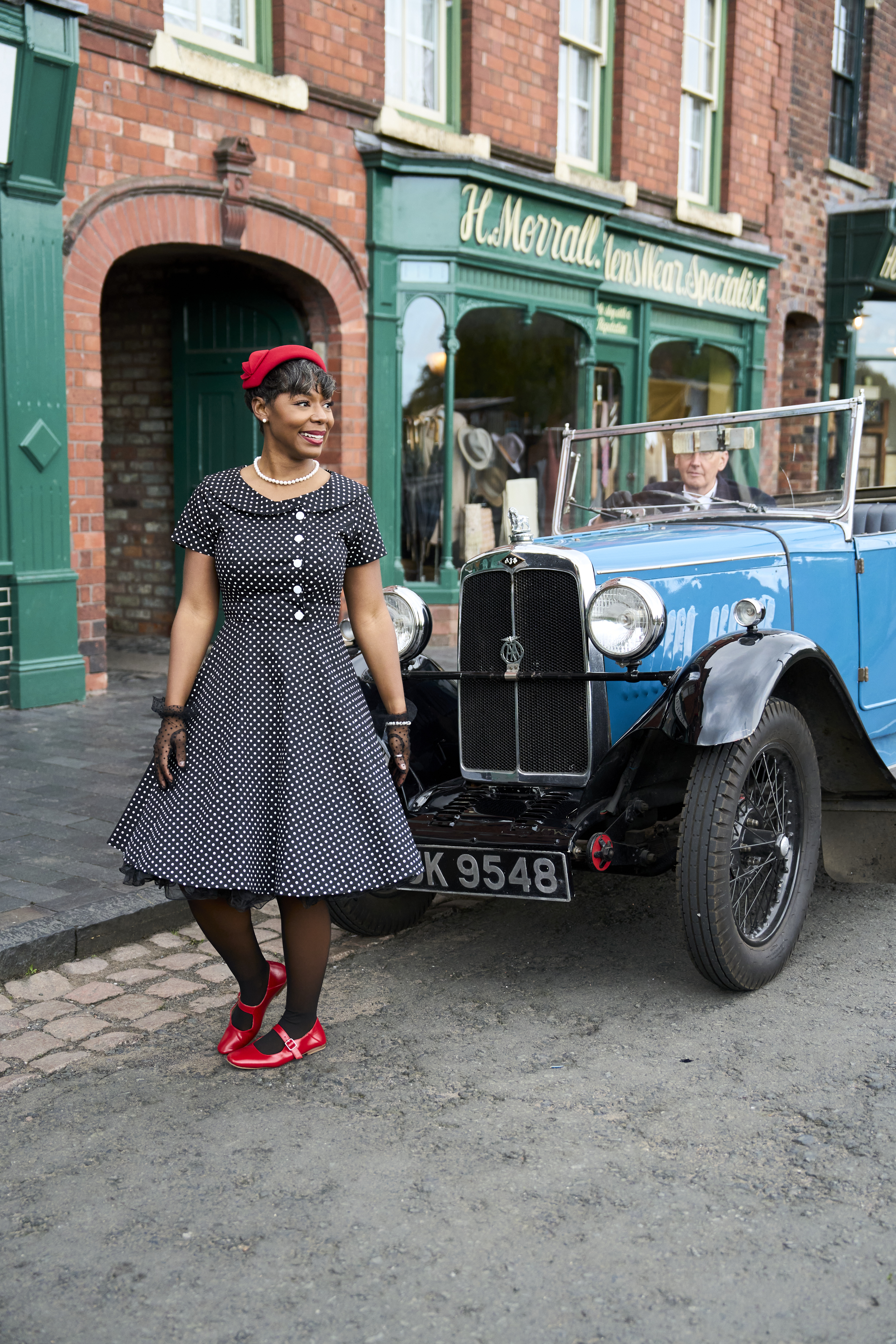 A woman dressed in vintage attire stands in front of a classic blue car parked on a street with period-style brick buildings in the background.