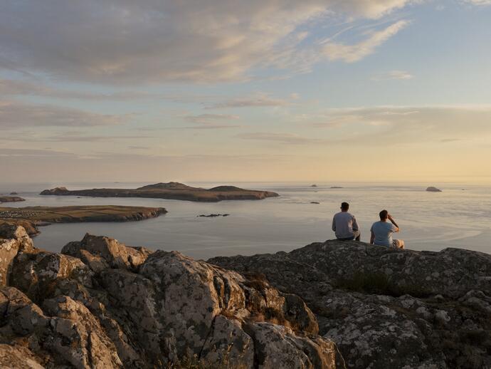 Dos hombres sentados en un acantilado mirando al mar al atardecer