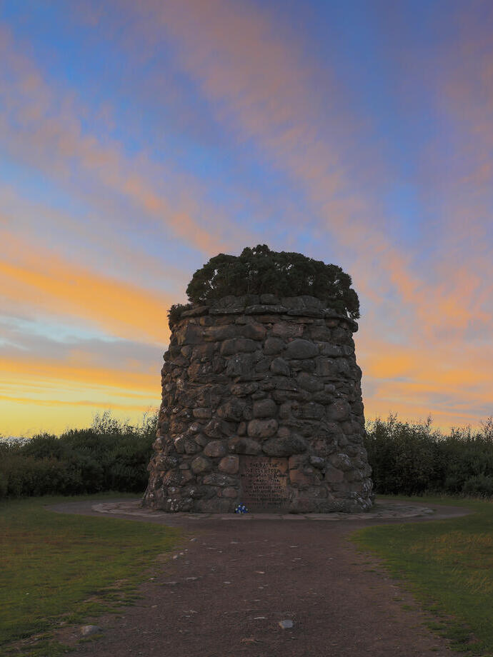 Ein kleiner Steinturm mit einer Gedenktafel, umgeben von Gras und Moor unter einem leuchtend blauen und orangefarbenen Himmel bei Sonnenuntergang.