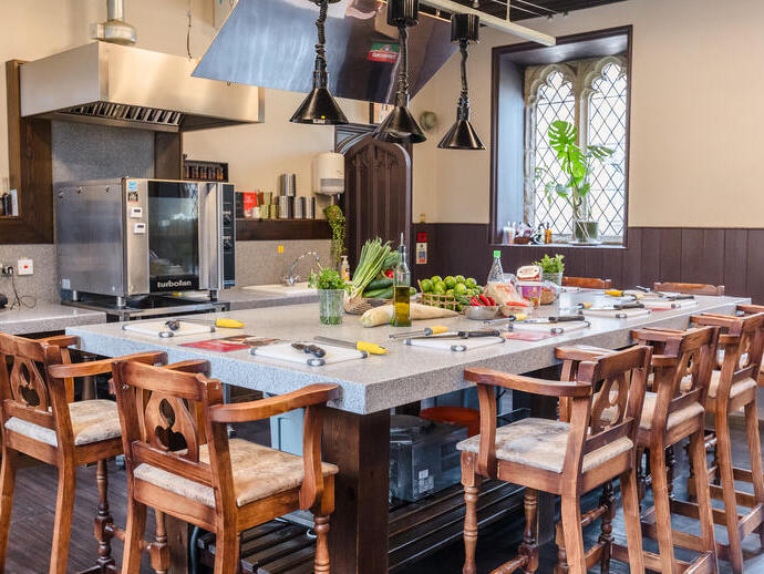 Modern kitchen with a central island, wooden chairs, prep stations, fresh ingredients, and a large oven. Bright window in the background.