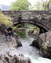 Deux hommes saluant depuis des rochers escarpés sous un pont de pierre enjambant une rivière étroite