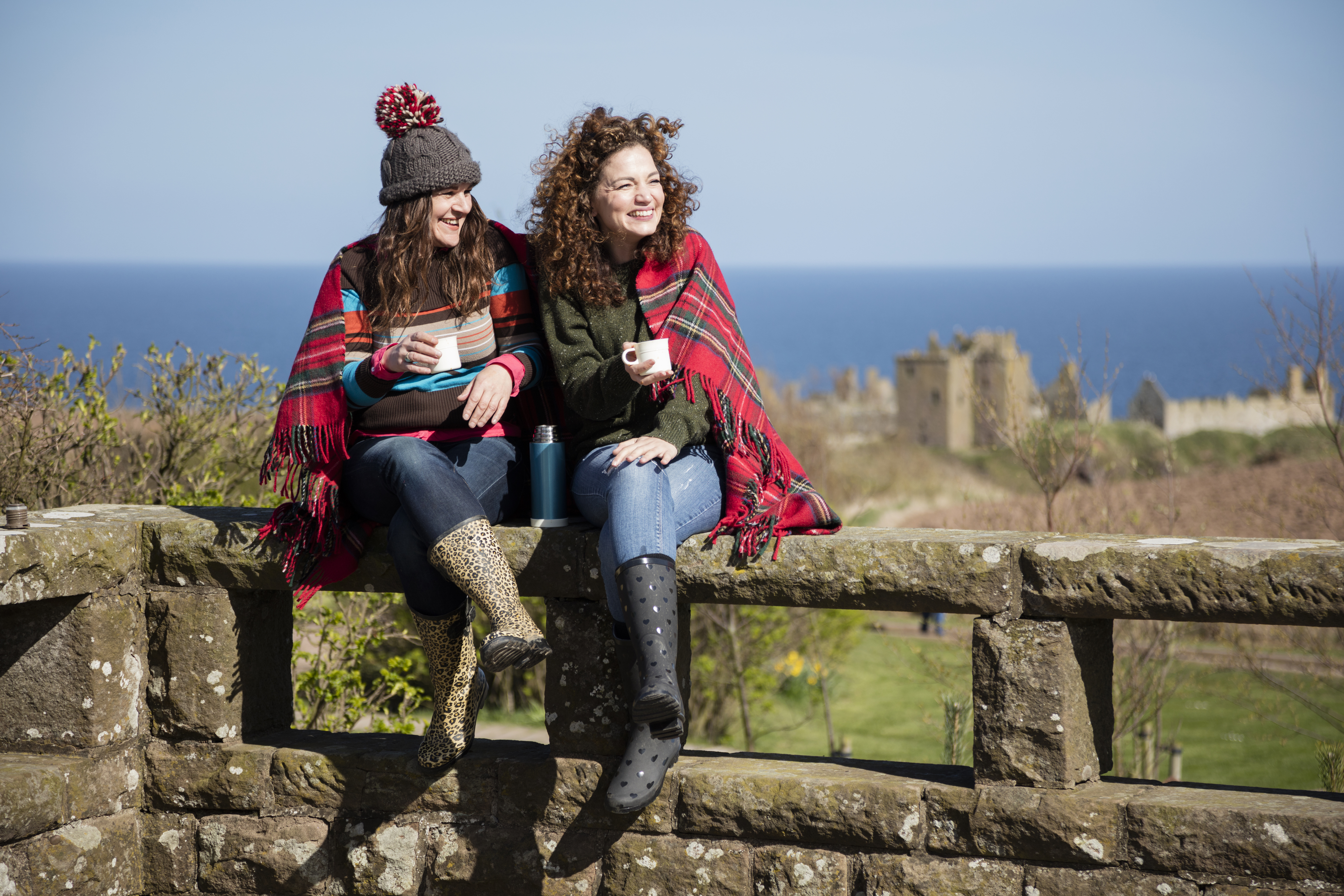 Two women with hot drinks, sitting on a stone wall near a castle