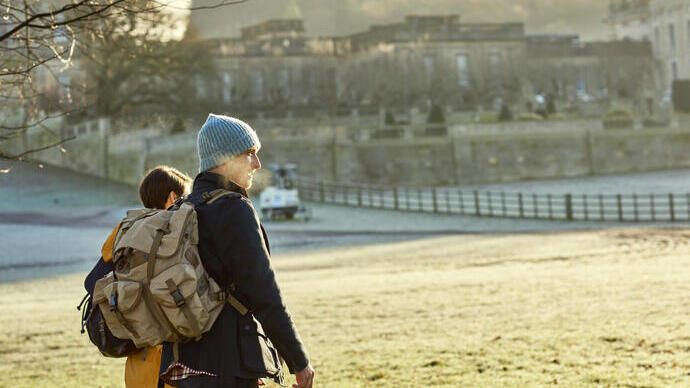 Two people with backpacks walk in a park on a cold morning, with a historic building and trees visible in the background.