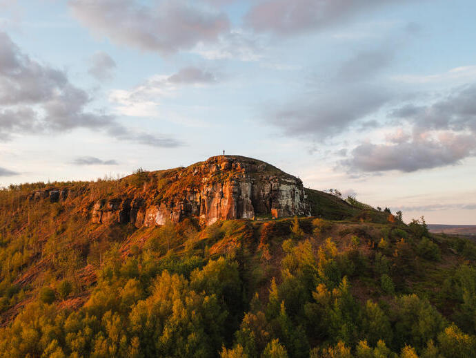 A lone person stands on a cliff top with forest below