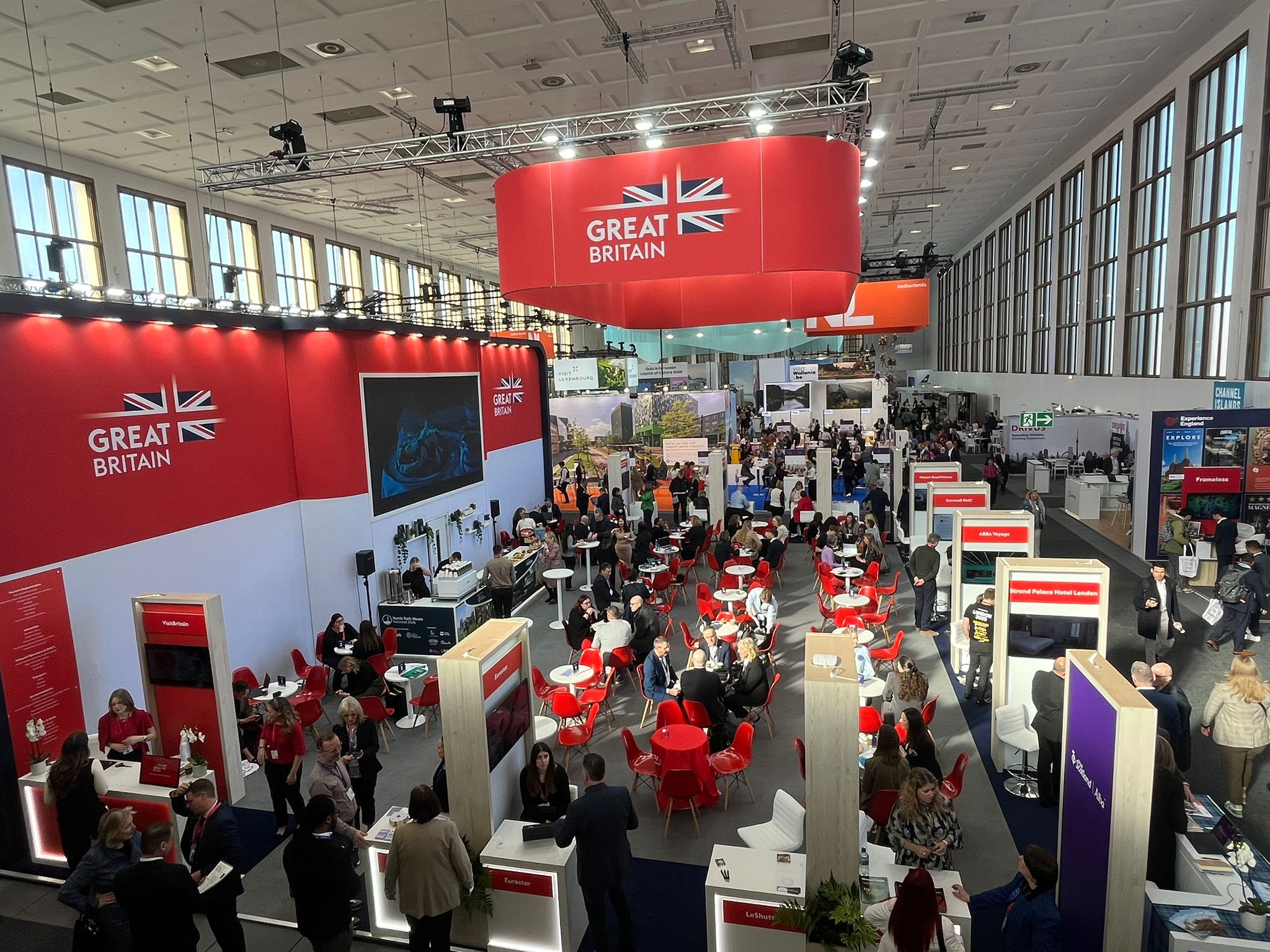 Busy trade show or exhibition hall with red and white Great Britain banners, people networking at tables, and various booths in a modern, bright venue.