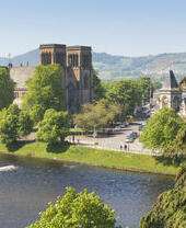 A cathedral with two towers overlooks a river on a sunny day, with trees in the foreground.