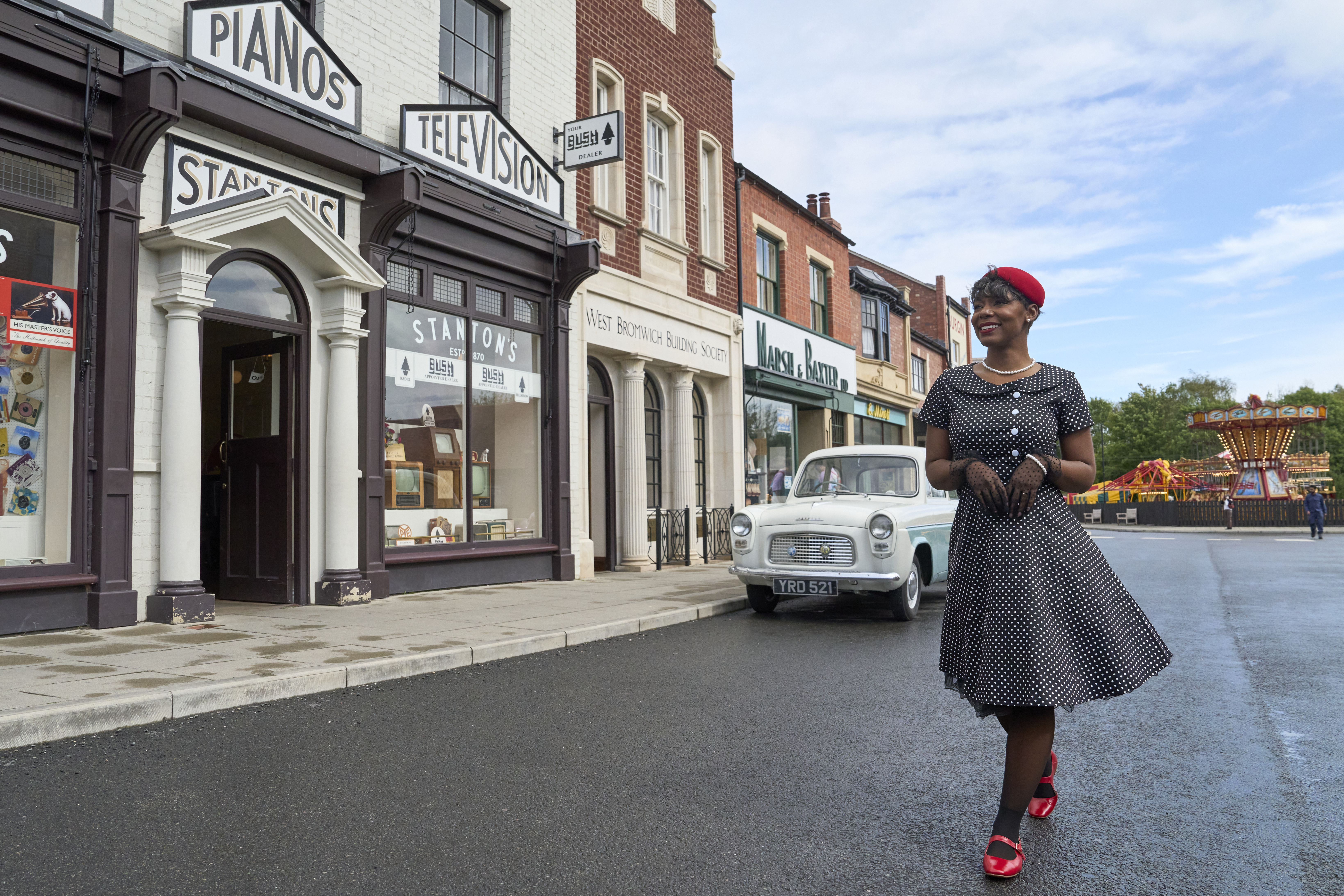 Woman in 1950s-style dress walks past vintage shops, classic car, and fairground ride on a recreated historic street scene.