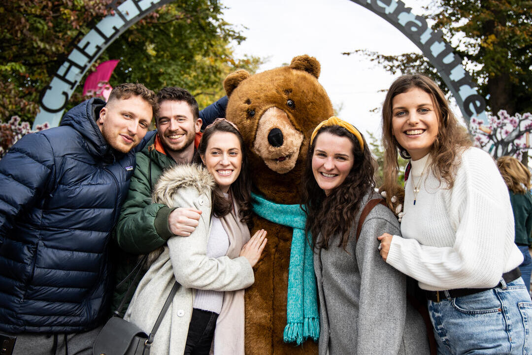People posing with big bear mascot under an arch sign reading Cheltenham Literature