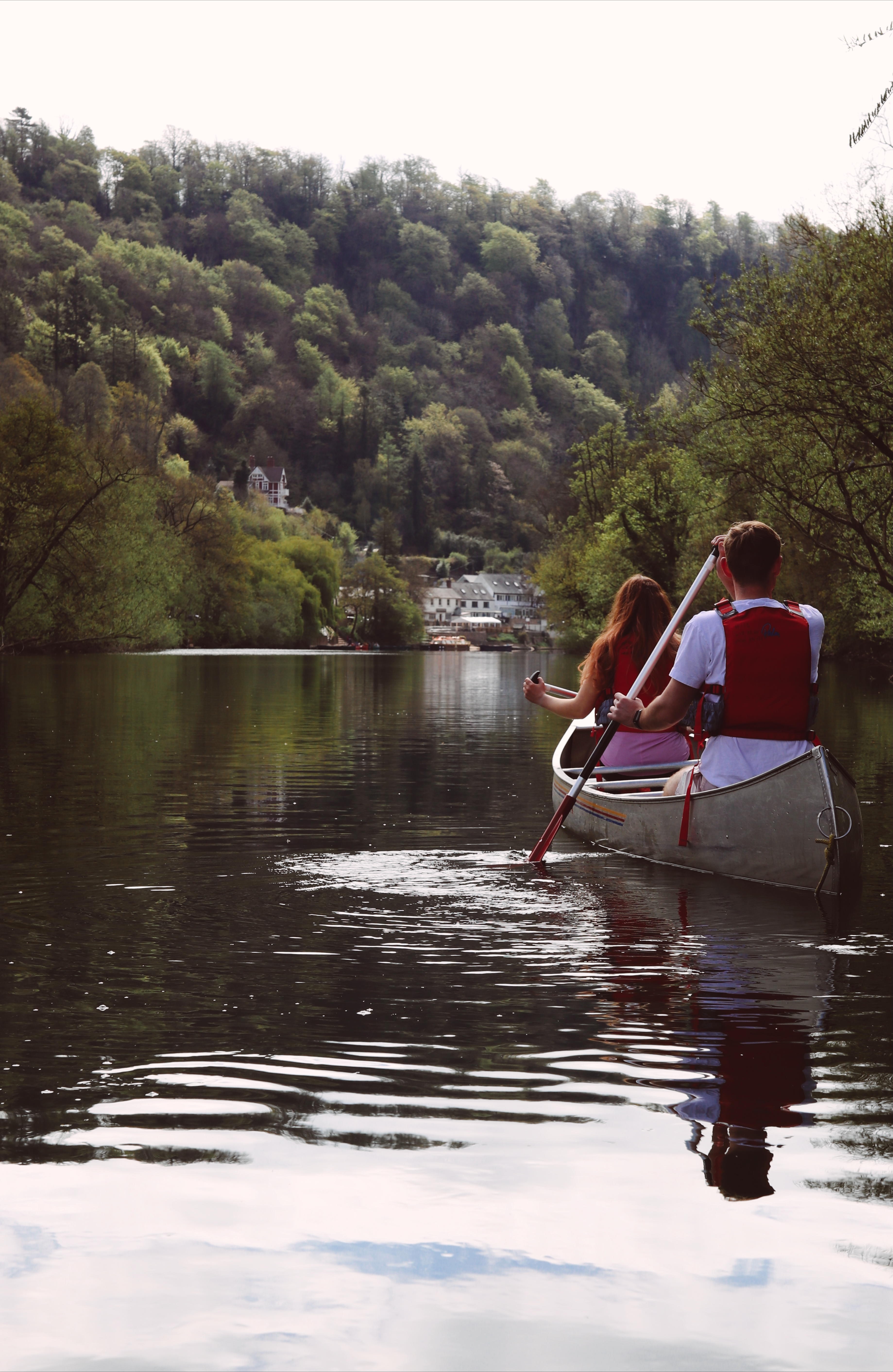 Two people paddling a canoe on a calm river surrounded by trees, with houses visible in the background.