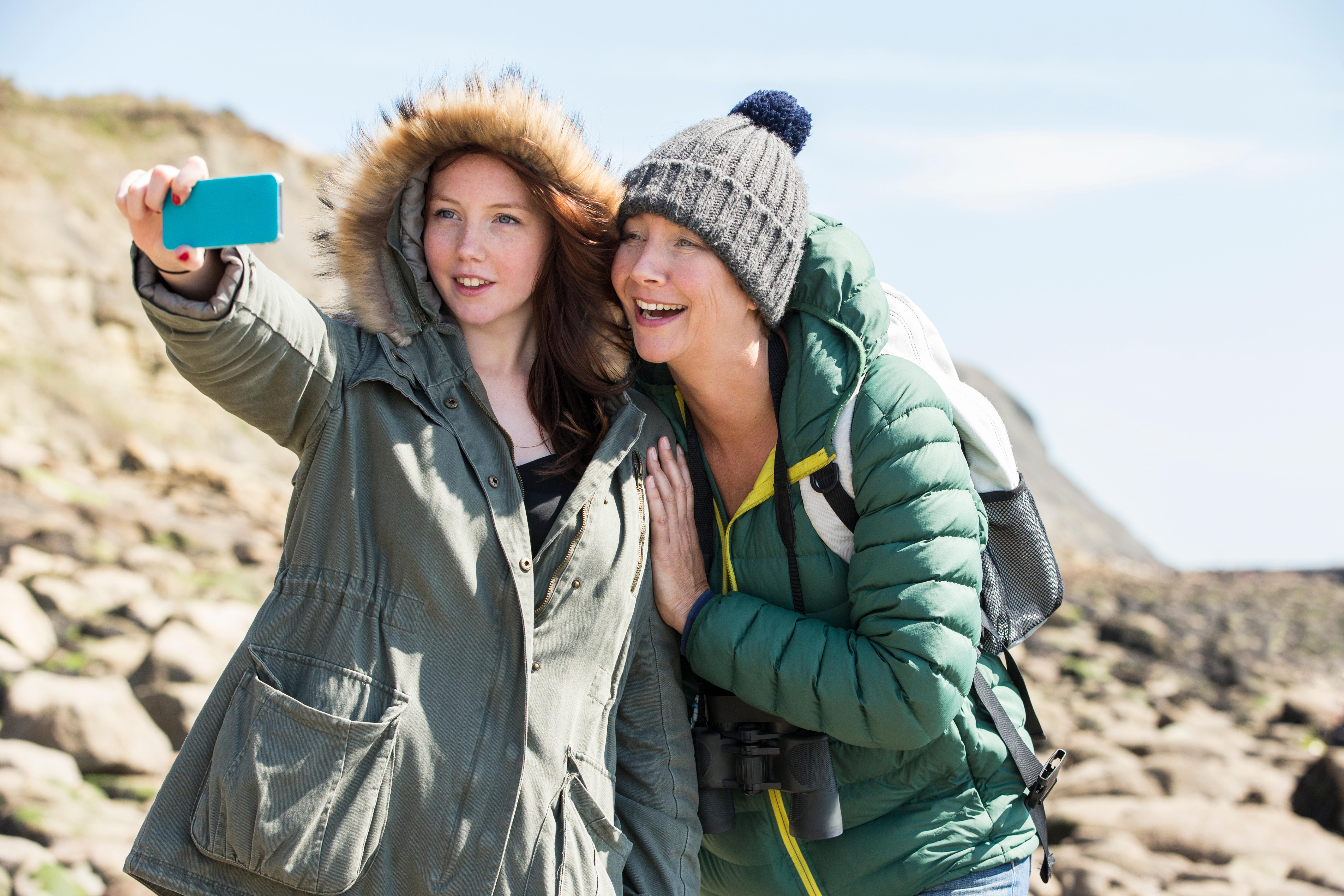 Mutter und Tochter machen ein Selfie an der Küste, sie tragen Hüte und Mäntel