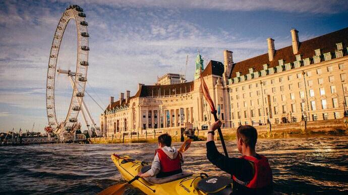 Zwei Personen fahren Kajak auf der Themse, mit dem London Eye und einem historischen Gebäude im Hintergrund.