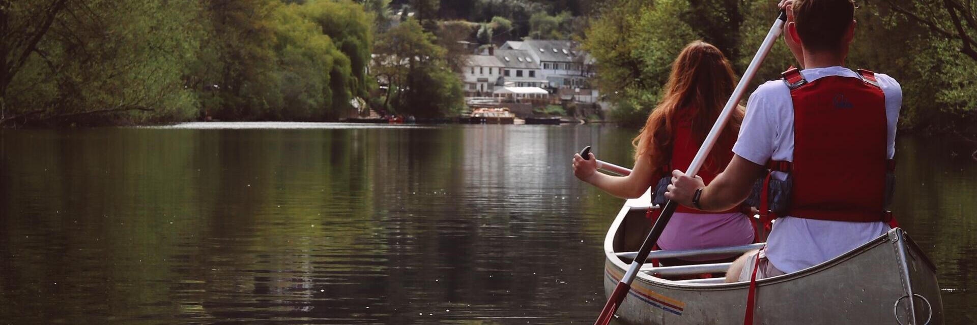 Due persone remano in una canoa su un fiume calmo circondato da alberi, con case sullo sfondo.