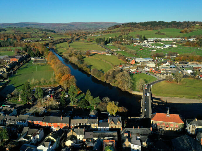 Aerial view of a small town with a river, a bridge, green fields, scattered buildings, and distant hills under a clear sky.