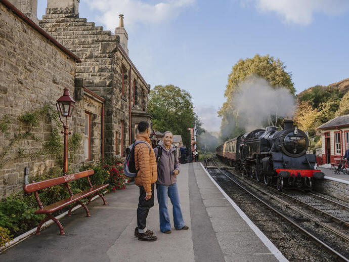 Dos personas conversan en un andén junto a un edificio de piedra antiguo y un tren de vapor sobre las vías. Al fondo hay árboles con follaje otoñal.
