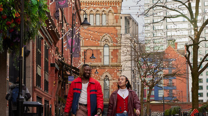 Two people walk along a brick street lined with cafes and string lights, with historic and modern buildings in the background.