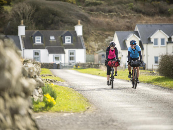 Two cyclists ride on a rural lane past stone walls and houses, with green grass and hills in the background.