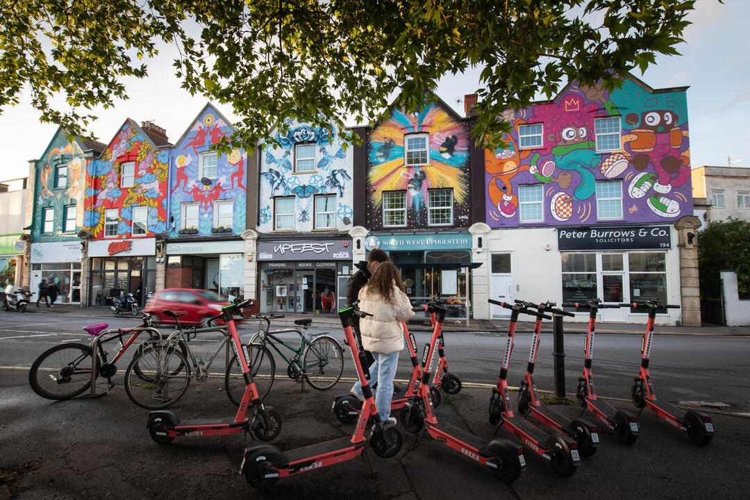 Two people standing by rental scooters in front of a row of houses painted on with colourful murals