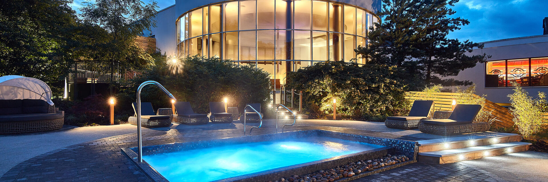 The outdoor spa area at dusk with a lit outdoor spa pool surrounded by loungers and the glass fronted circular spa building in the background. 