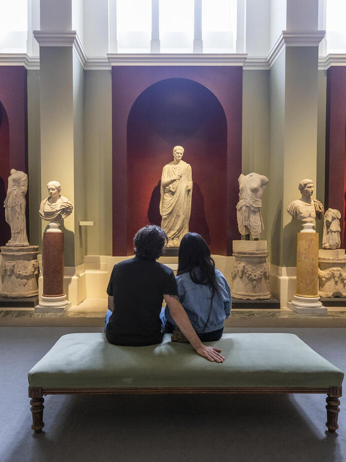 A woman and a man sit in a museum looking at Greek and Roman sculptures