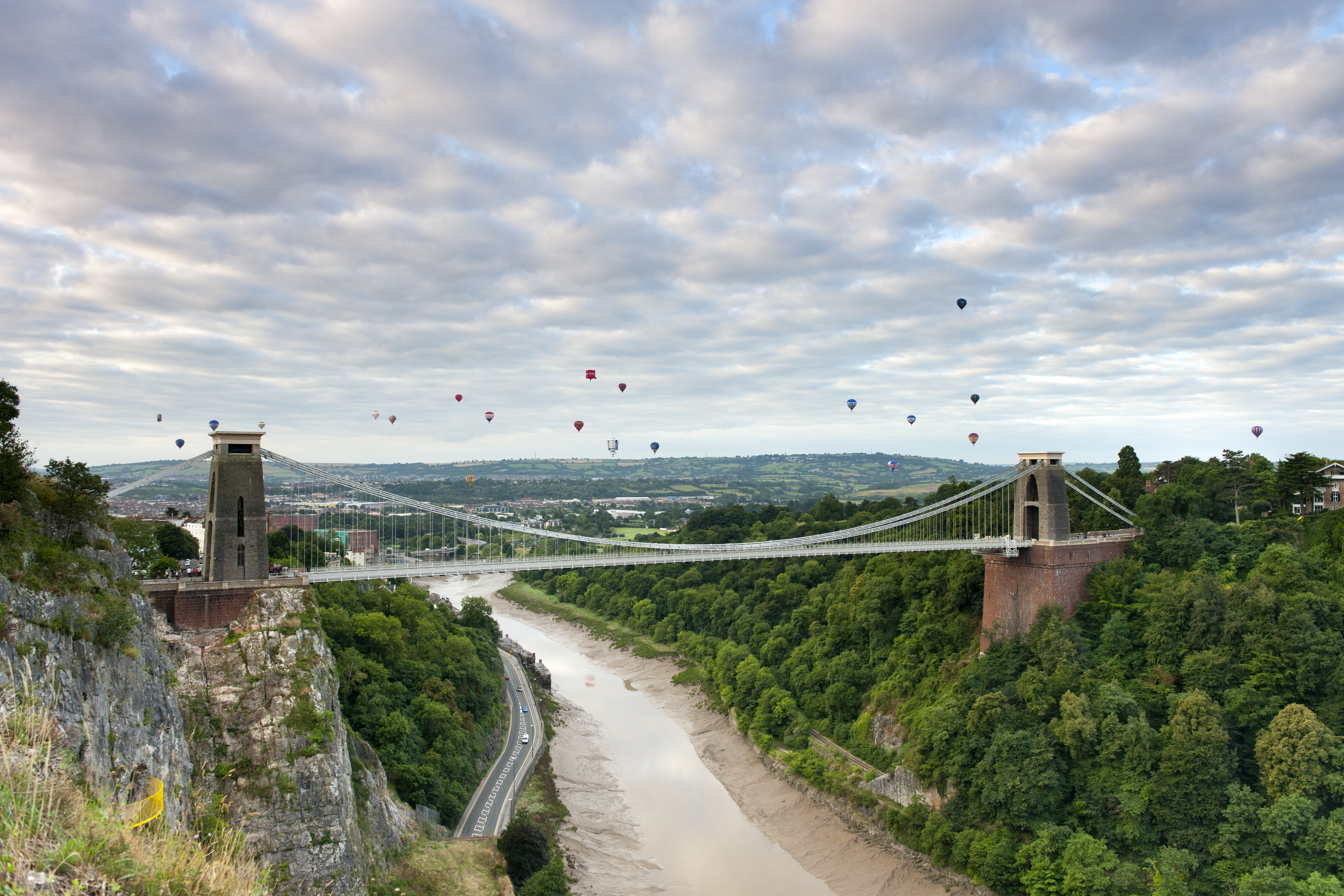 Hot air balloons floating above a river and a suspension bridge.