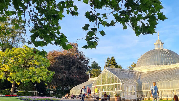 Large domed glass house building in the background in the sunshine, with people outside walking though the park, flower beds in foreground.