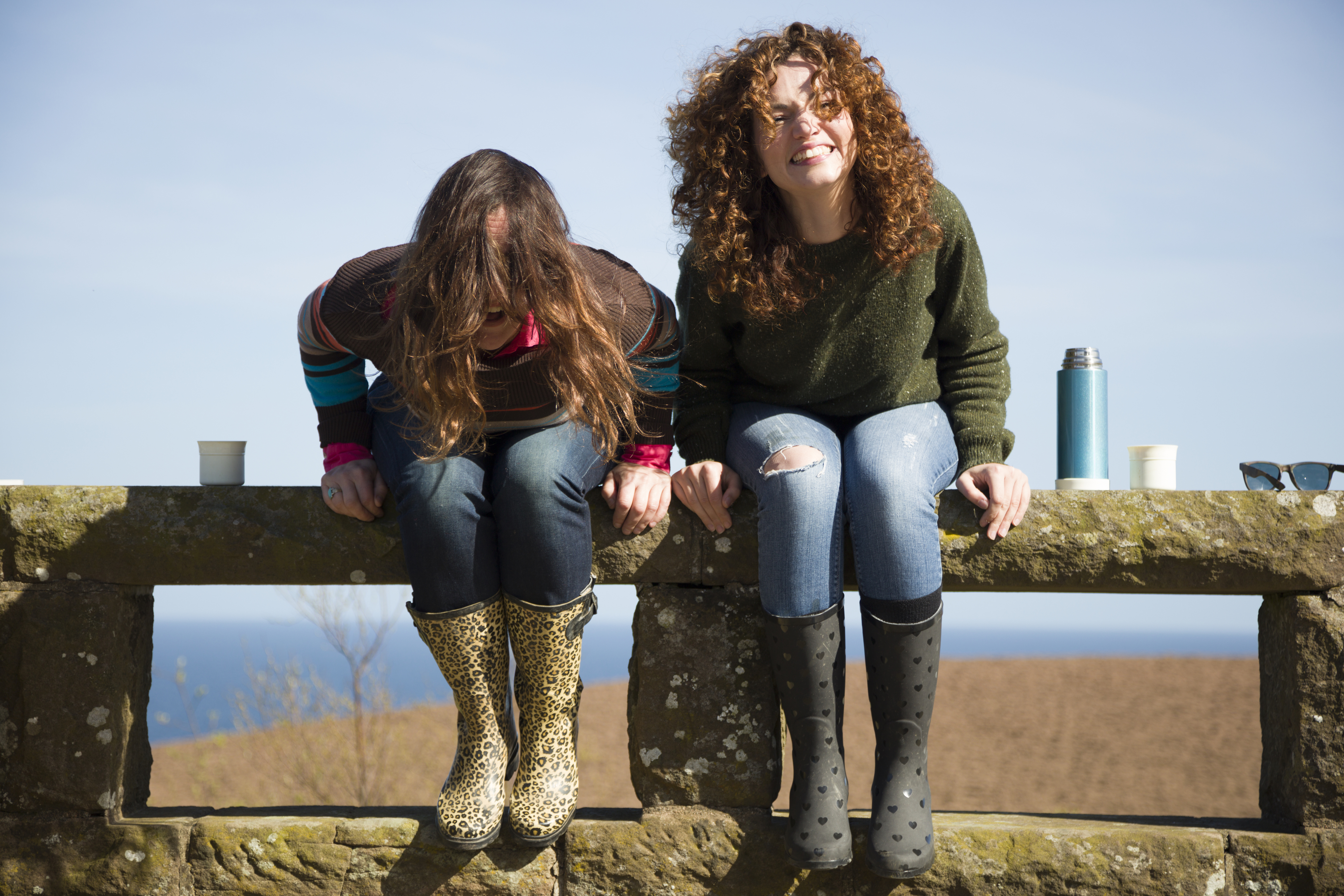 Two women laughing, sat on a stone fence