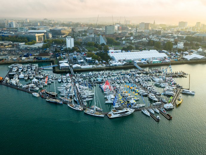 Aerial view of a marina filled with boats and yachts by the waterfront. Modern cityscape and event tents are visible in the background.