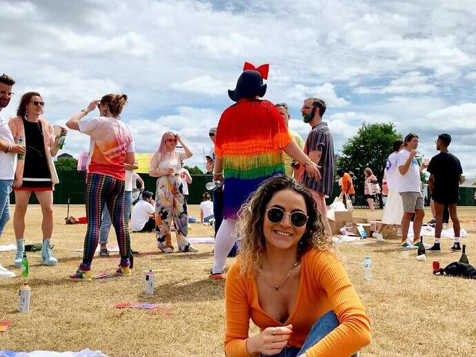 Mujer con gafas de sol sonriendo, sentada en la hierba en un festival con gente al fondo