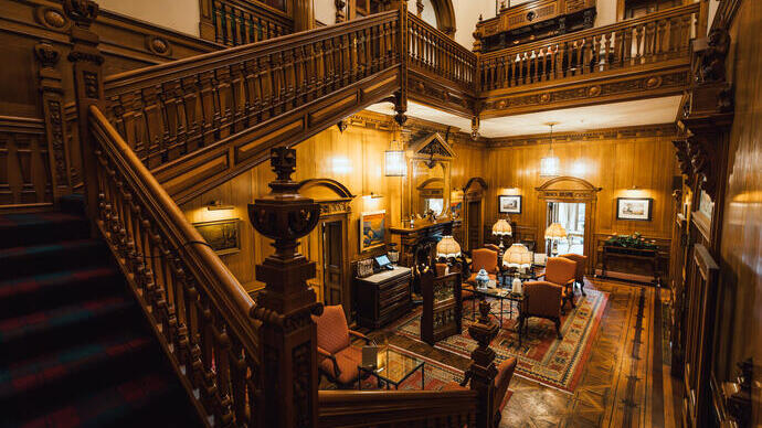Opulent wood-paneled interior with ornate staircase, classic furniture, lamps, carpets and framed pictures, evoking a grand historic home.