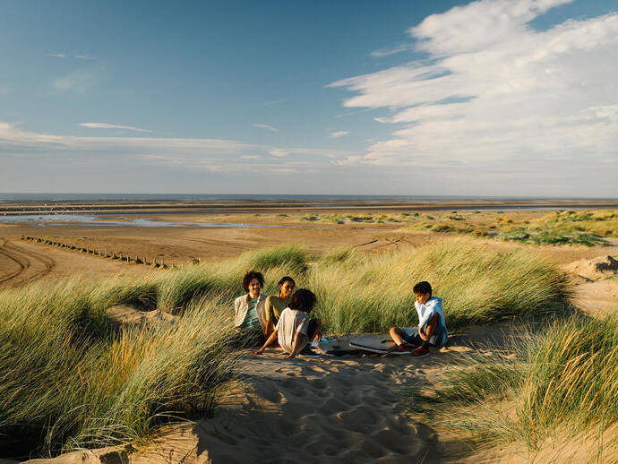 Four people sit on sand dunes among tall grass, with a wide sandy beach and tidal flats extending to the horizon under a blue sky.
