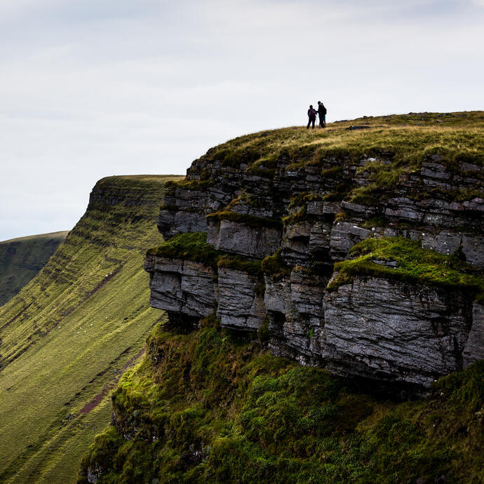 Two people stand atop a grassy cliff with dramatic rocky edges, overlooking rolling green hills under a cloudy sky.