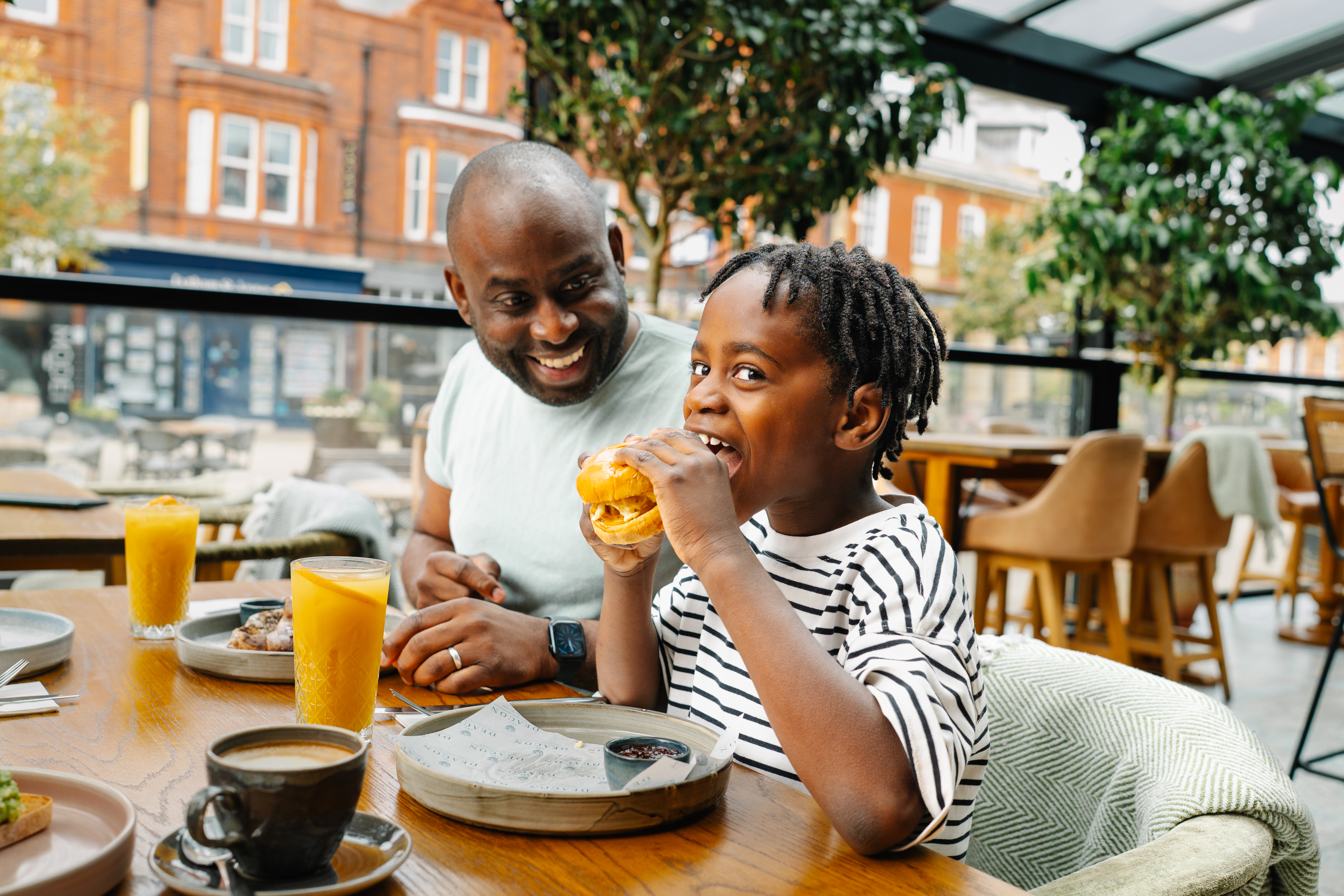 Child eating a burger and adult smiling at an outdoor restaurant table with drinks, plants, and red brick buildings in the background.