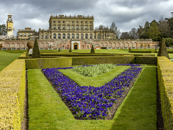 Beautiful manicured flower gardens and sculptured hedges in front of a large mansion.