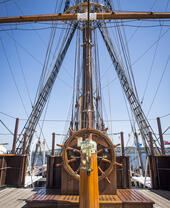 Wooden ship's wheel and rigging on the deck of a historic sailing vessel, with blue sky and seaside background.