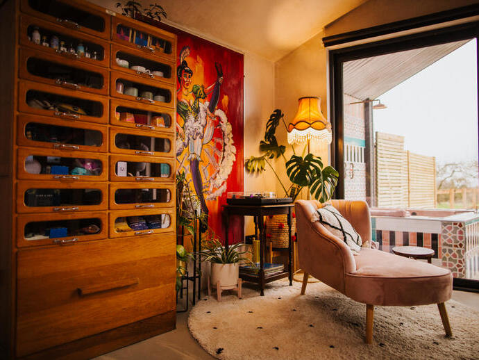 A pale pink chaise lounge on a rug lookng out of floor to ceiling windows. A tall wooden dresser to the left and vibrant artwork on the walls
