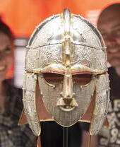 Two people admire an ornate Anglo-Saxon helmet in a museum display with a bright orange backdrop.