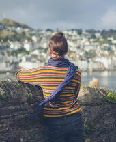 A young woman is standing by a stone wall in a village and is looking at the boats moored in the estuary