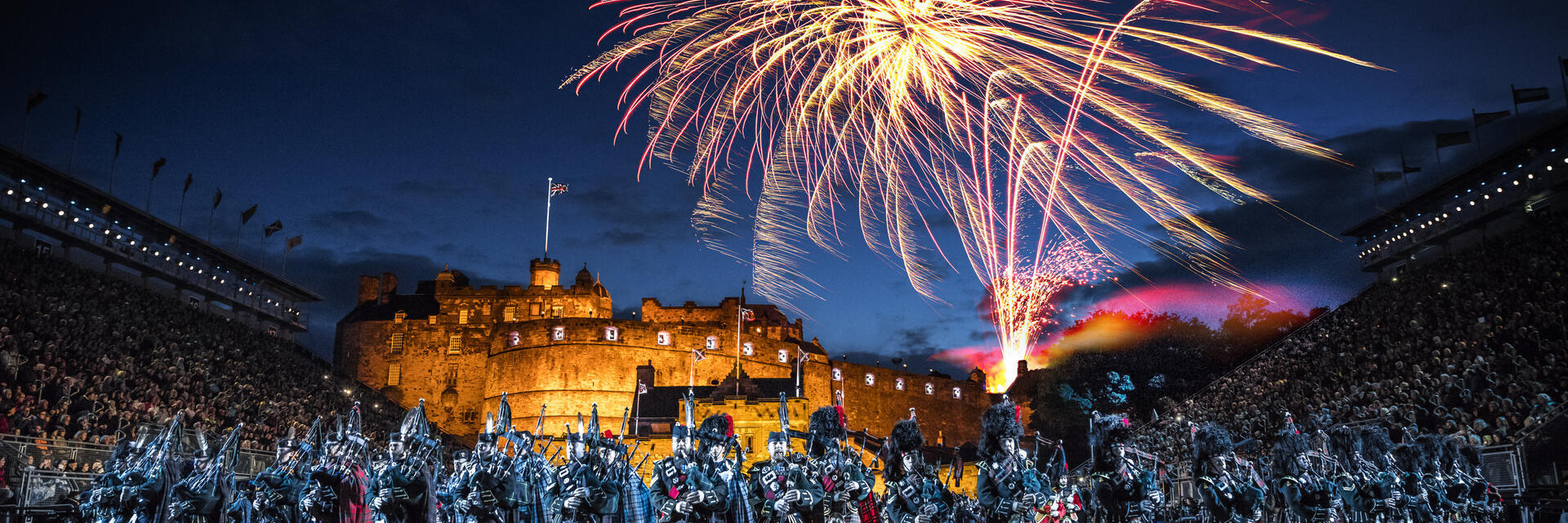 The arena at the Edinburgh Military Tattoo during a performance of the military event, parade ground and packed spectator seating. A light show projecting onto the castle walls. Marching band with a leading conductor, and massed pipers playing the bagpipes. Fireworks exploding in the night sky.