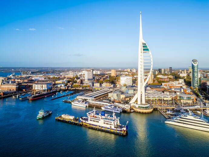 A waterfront cityscape featuring a tall, modern tower, ferries, yachts, and urban buildings under a clear blue sky.