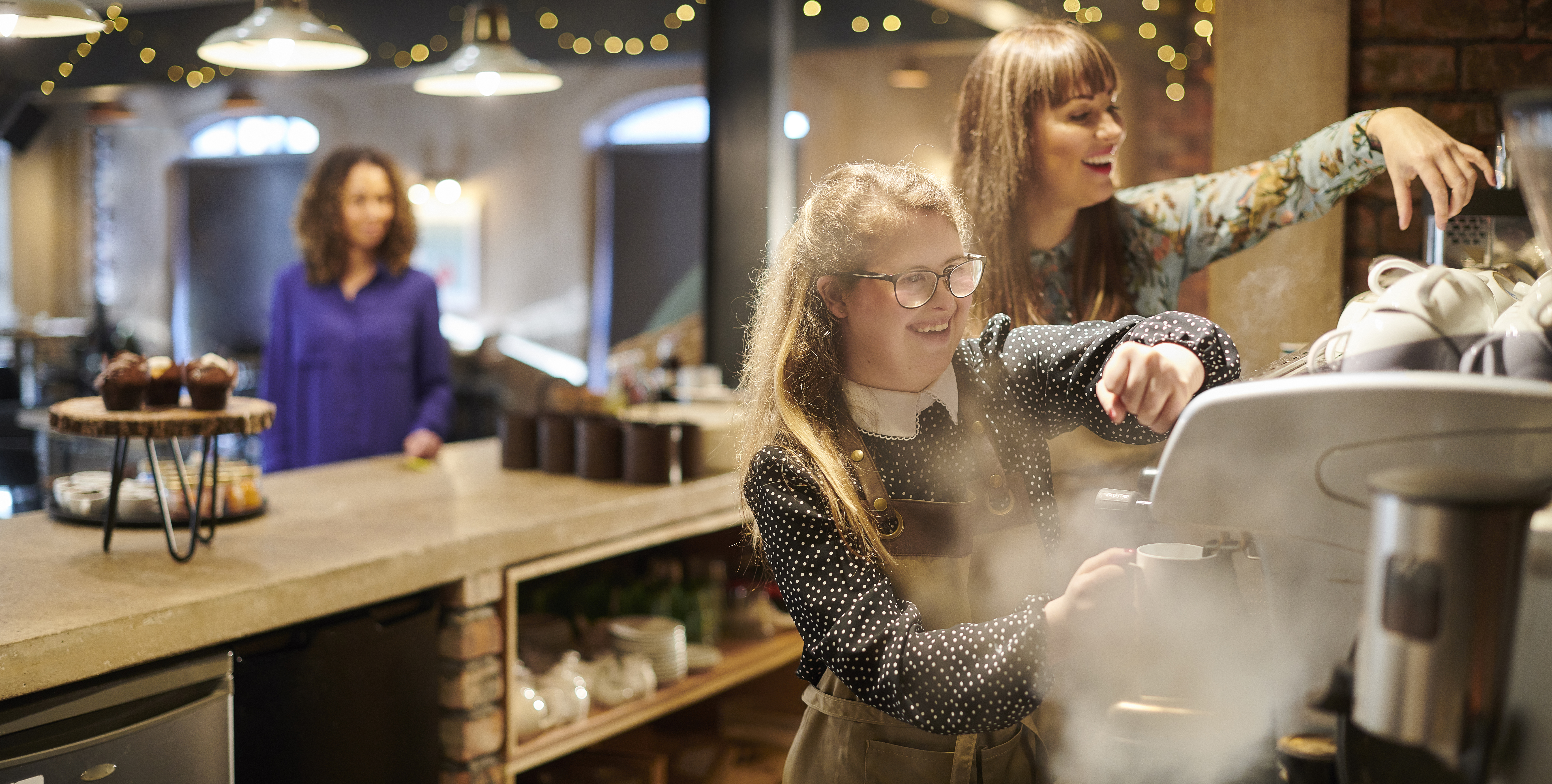 A young woman working the coffee machine with colleague assisting in cafe