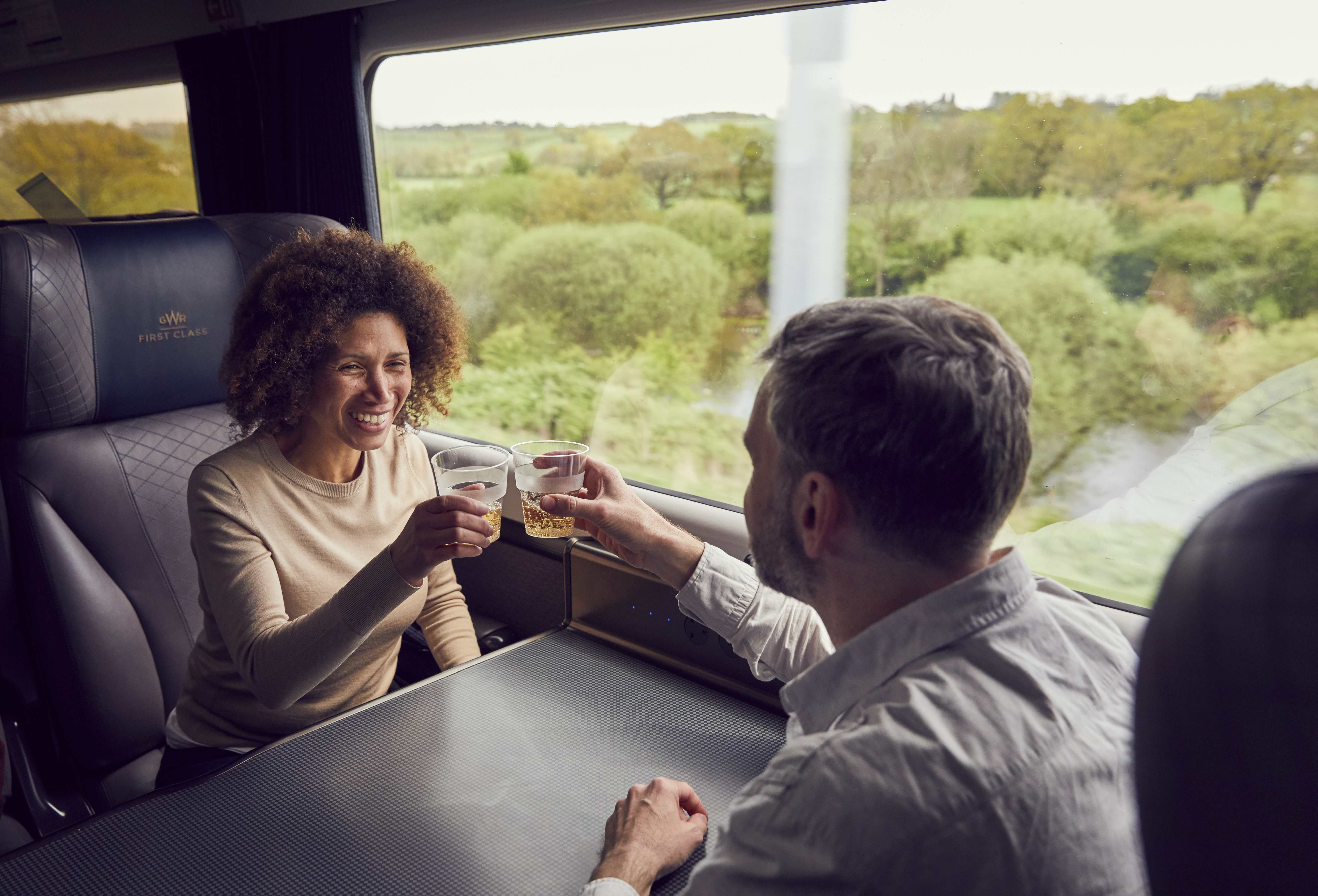 A couple enjoying a drink while travelling by train from Bath, sitting at a table, toasting