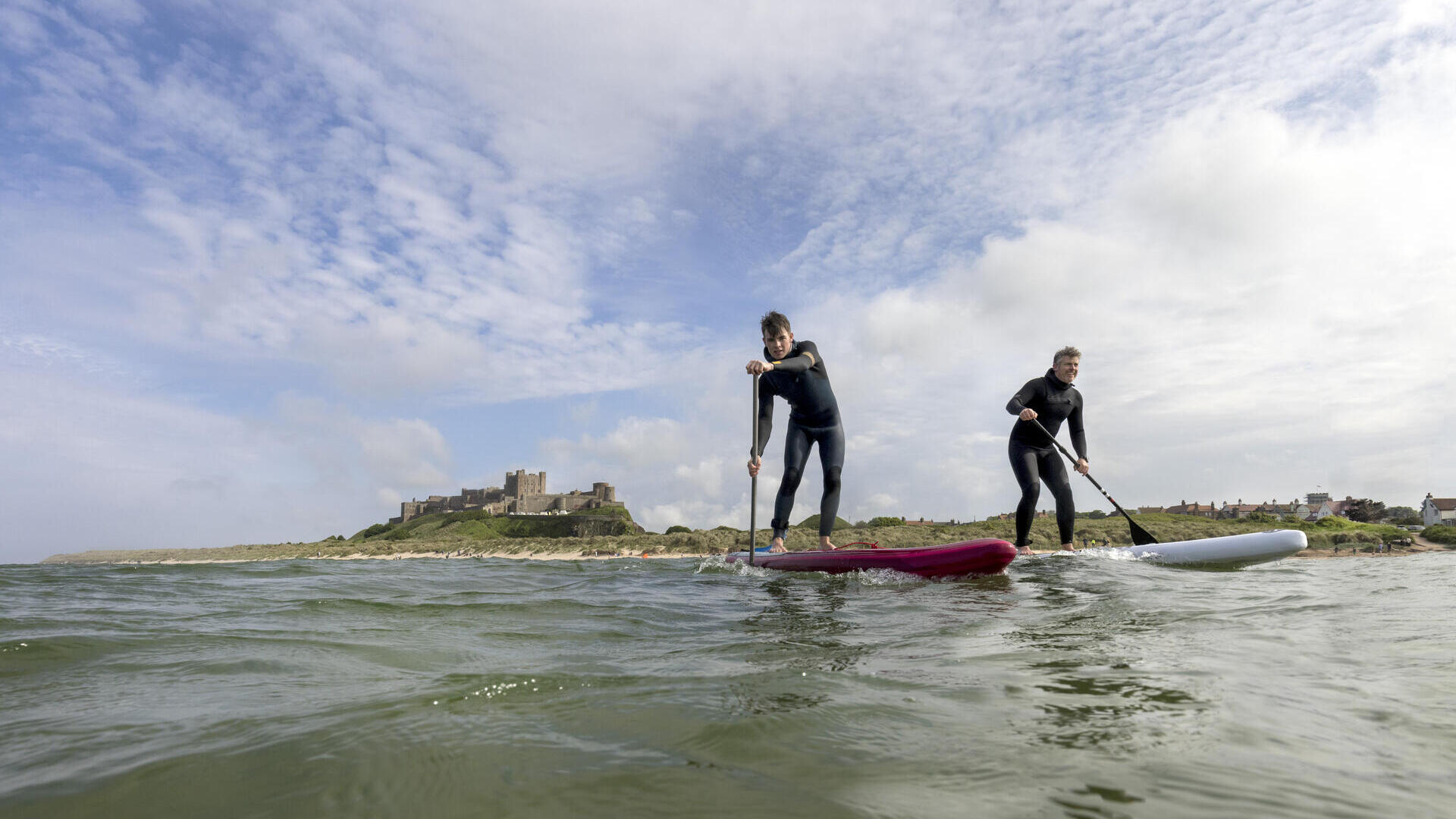 Deux hommes font du paddleboard dans la mer avec un château historique en arrière-plan.