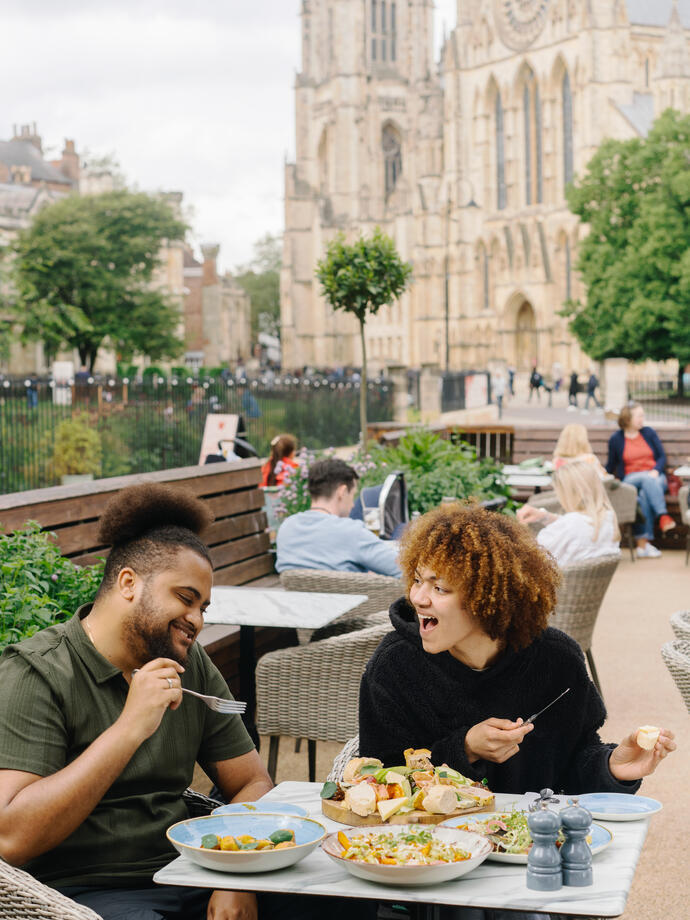 A man and a woman eat lunch outside a cafe with a cathedral in the background