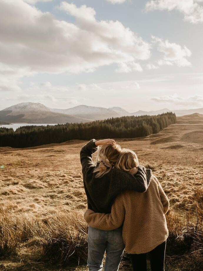 Zwei Freunde umarmen sich und blicken über das Tal auf einen See und die Berge