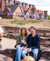 Two people sitting and listening to a tour guide on a Roman themed tour of Chester