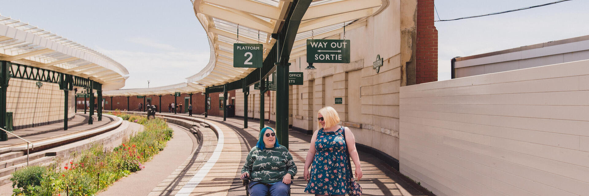 Una mujer camina con otra mujer que está sentada en una silla de ruedas en una antigua estación de tren.