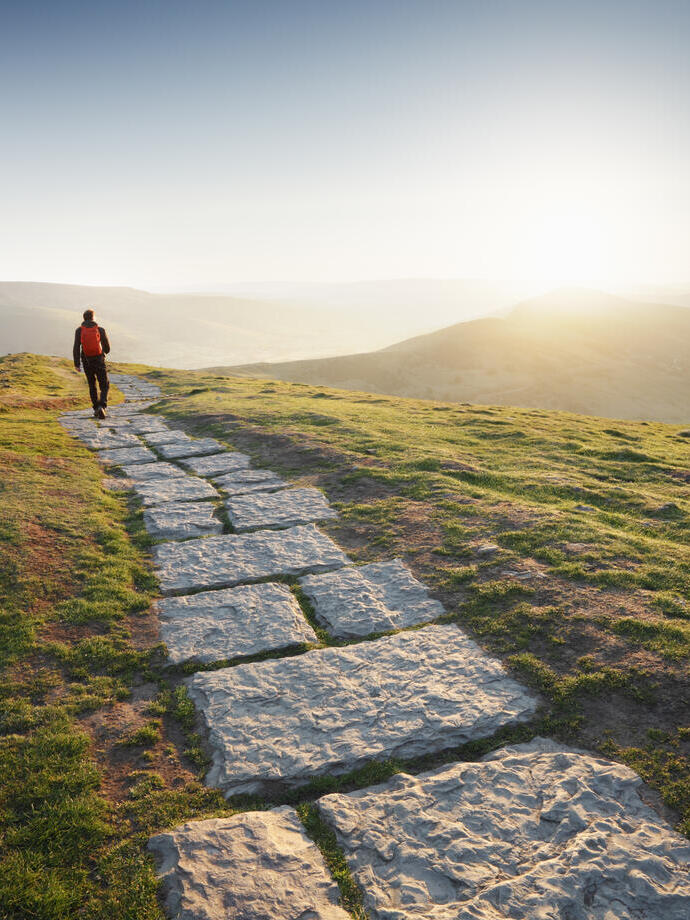Man in red jacket walking away into the sun at the top of a mountain path