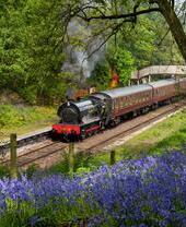 Un train à vapeur d'où s'échappe beaucoup de vapeur, tirant au moins trois wagons d'époque, se prépare à quitter une petite gare ferroviaire traditionnelle.
