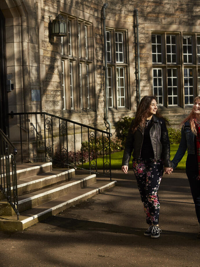 Two women holding hands whilst walking past a university building.